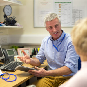 A male doctor sits face to face with an elderly woman. They are in the doctors office . The focus is on the doctor who sits at his desk holding a digital tablet and smiling at the woman. The woman face cannot be seen.